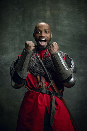 Half-length portrait of dark skinned man, medieval warrior or knight wearing armour shouting as winner isolated over dark vintage background. Comparison of eras, historyの写真素材