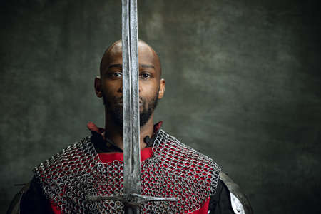 Vintage portrait of dark skinned medieval warrior wearing chain mail holding big sword isolated over dark vintage background. Comparison of eras, history. Side viewの写真素材