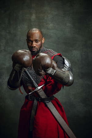 Vintage style portrait of brutal dark skinned man, medieval warrior or knight wearing armour in boxing gloves isolated over dark background. Comparison of eras, history, sportの写真素材