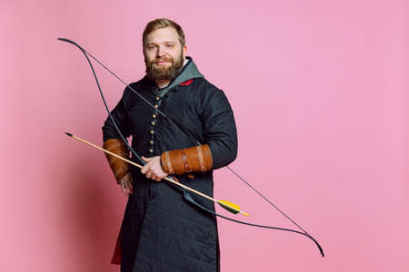 Studio shot of young smiling bearded man in image of medieval warrior, archery in chain armor...