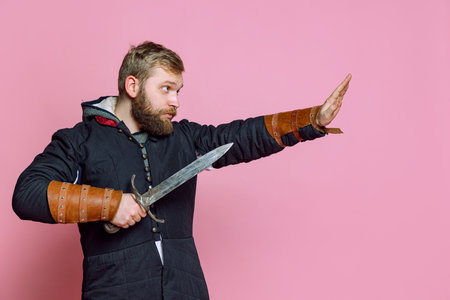 Portrait of brave man, medieval warrior or archer in armor with sword rising hand showing stop gesture isolated over pink studio backgroundの写真素材