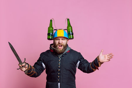 Half-length portrait of young bearded man in image of medieval warrior, archer in beer helmet with sword isolated over pink studio backgroundの写真素材