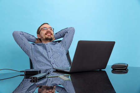 Portrait of happy looking man, computer support specialist, programmer sitting in front of laptop isolated over blue backgroundの写真素材