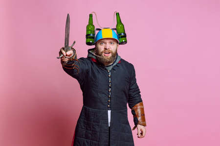 Half-length portrait of young bearded man in image of medieval warrior, archer in beer helmet with big knife isolated over pink studio background. Beer festival conceptの写真素材