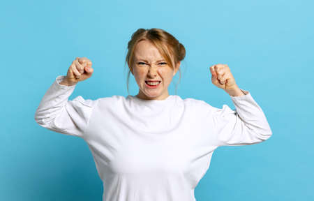 Angry young caucasian girl wearing white pullover clenches fists isolated on blue background....