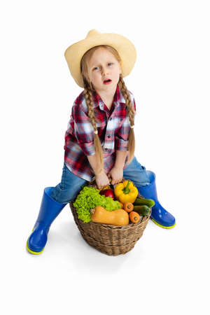 Studio shot of cute little girl, emotive kid in image of farmer, gardener holding big basket of vegetables isolated on white background. Model in plaid shirt, jeans and gumbootsの写真素材