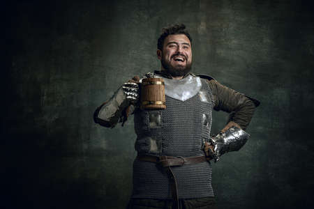 Happy medieval warrior or knight with dirty wounded face holding big mug of beer isolated over dark vintage background. Comparison of eras, historyの写真素材