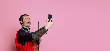 Comic portrait of medieval warrior or knight wearing wearing armor clothing using phone isolated over pink background. Comparison of eras, historyの写真素材