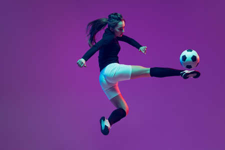 In action. One sportive girl, female soccer player training with football ball isolated on purple studio background in neon light. Sport, action, motion, fitnessの写真素材