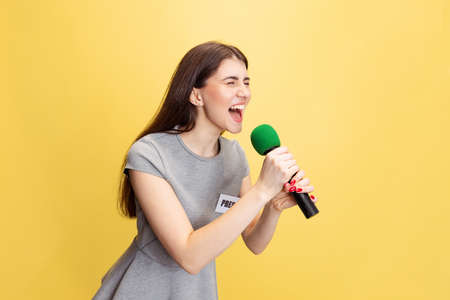 Emotional young girl, female reporter holding reporter microphone isolated on yellow studio background. Concept of social media, press, news, informationの写真素材