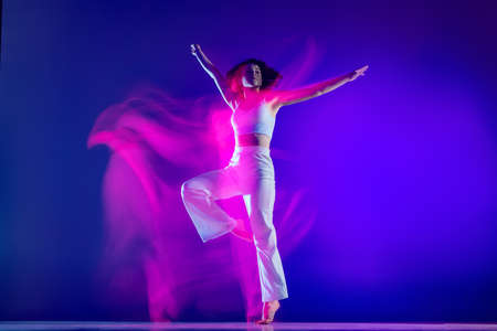 Dynamic portrait of beautiful girl, hip-hop dancer in white outfit dancing hip hop isolated on blue background in pink neon light. Youth culture, style and fashion, action.の写真素材