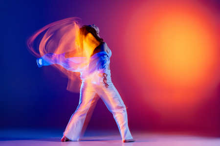Image of flexible young girl, hip-hop dancer in white outfit dancing hip hop isolated on blue background in yellow neon light. Youth culture, style and fashion, action.の写真素材