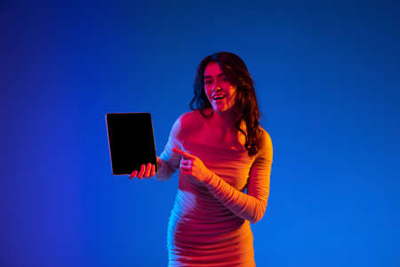 Portrait of young emotional girl, student with long dark hair using gadget isolated on dark blue background in neon light. Concept of emotionsの写真素材