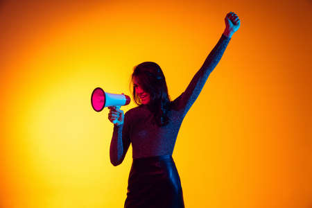 Beautiful young excited girl, student shouting at megaphone isolated on gradient yellow background in neon light. Concept of emotions, youthの写真素材