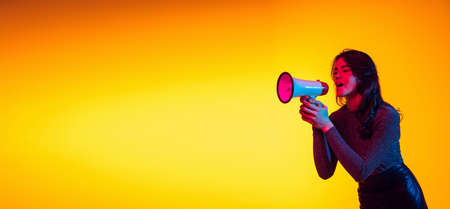 Beautiful young excited girl, student shouting at megaphone isolated on gradient yellow background in neon light. Concept of emotions, youthの写真素材