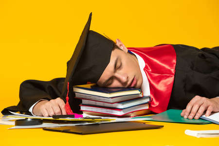 Portrait of young man, student in graduation cap and gown sleeping on books isolated over yellow studio backgroundの写真素材