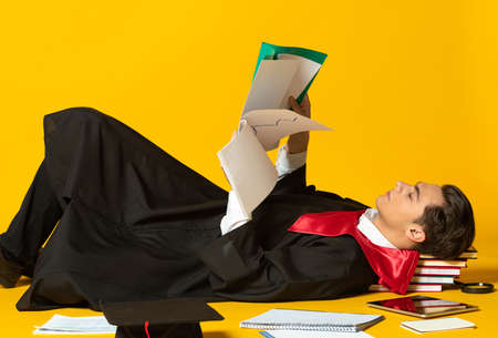 Portrait of young man, student in graduation hown lying on floor and reading papers isolated oer yellow studio backgroundの写真素材