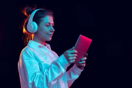 Portrait of young beautiful happy girl in headphones using tablet isolated on orange background in neon light, filter. Concept of emotions, musicの写真素材