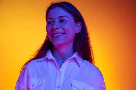 Smile. Portrait of young beautiful emotive girl in white shirt isolated on orange background in neon...