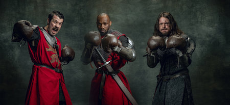 Portraits of medieval warriors or knights with dirty wounded face in boxing gloves isolated over dark background. Comparison of eras, history, sportの写真素材