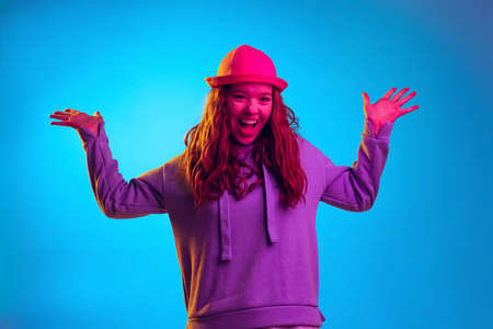 Portrait of young stylish emotive girl in hoodie cheerfully posing isolated over blue studio background in pink neon lightの写真素材