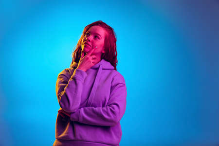 Half-length portrait of young emotive girl, thoughtfully looking away isolated over blue studio background in pink neon lightの写真素材