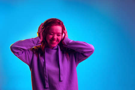 Portrait of young emotive girl cheerfully listening to music in headphones isolated over blue studio background in pink neon lightの写真素材