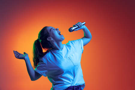 Portrait of young girl emotively singing in microphone, posing isolated over gradient red background...
