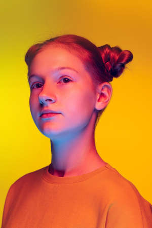 Portrait of beautiful young girl in orange sweatshirt looking away, posing isolated over yellow background in neon lightの写真素材