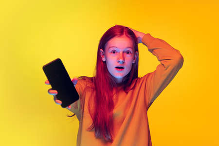 Portrait of young emotive girl having online video call on phone isolated over yellow studio...