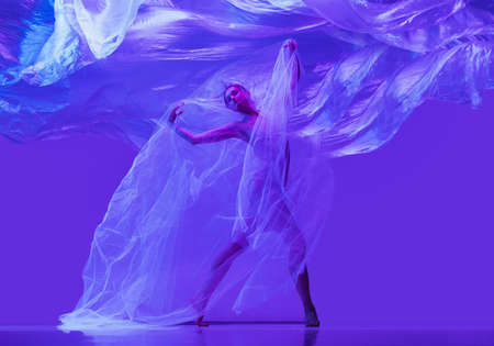 Portrait of beautiful flexible woman, ballerina dancing with cloth on purple studio background in neon. Fashion, style, art, beautyの写真素材