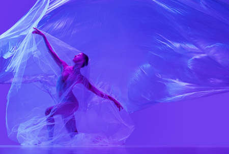 Portrait of beautiful flexible woman, ballerina dancing with cloth on purple studio background in neon. Fashion, style, art, beautyの写真素材