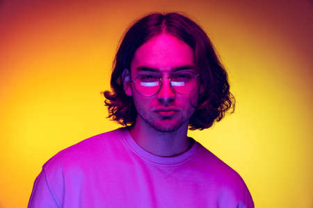 Portrait of young man, student in casual clothes isolated on orange background in purple neon light. Concept of emotions, beauty, fashionの写真素材