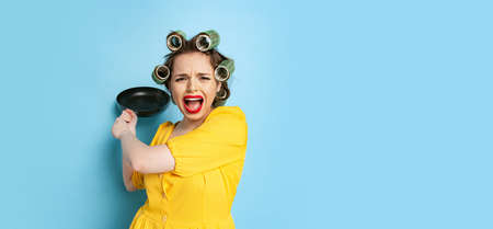 Comic portrait of young emotional girl, housewife with curlers on her head holding pan isolated on blue studio background. Emotions, female rights, beauty, familyの写真素材