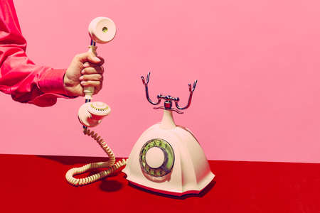 Pop art photography. Retro objects, gadgets. Female hand holding handset of vintage phone isolated on pink and red background. Vintage, retro fashion styleの写真素材