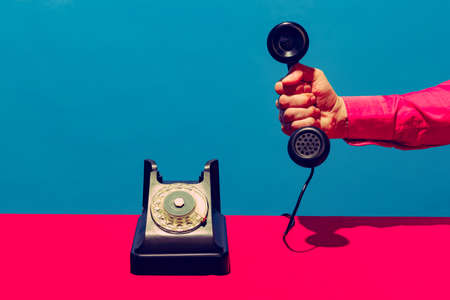 Retro objects, gadgets. Female hand holding handset of vintage phone isolated on blue and pink background. Vintage, retro fashion style. Pop art photography.の写真素材