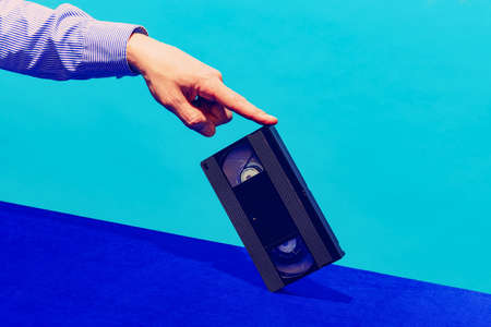 Pop art photography. Female hand holding cassette for video recorder isolated on blue background. Vintage, retro fashion styleの写真素材