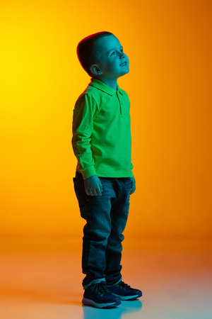 Full-length portrait of charming little boy posing isolated on yellow studio backgroud in neon light. Concept of child emotions, facial expression, action, childhoodの写真素材