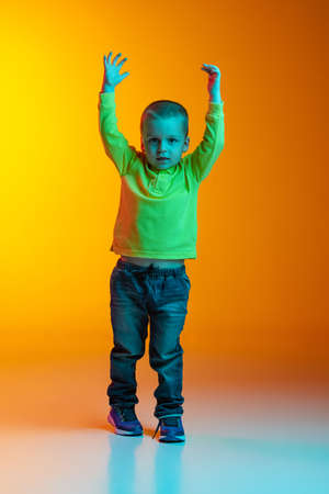 Full-length portrait of charming little boy posing isolated on yellow studio backgroud in neon light. Concept of child emotions, facial expression, action, childhoodの写真素材