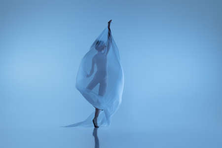 Portrait of flexible woman, graceful ballerina dancing with fabric, cloth isolated on blue studio background. Grace, art, beauty concept. Weightless, tenderness.の写真素材