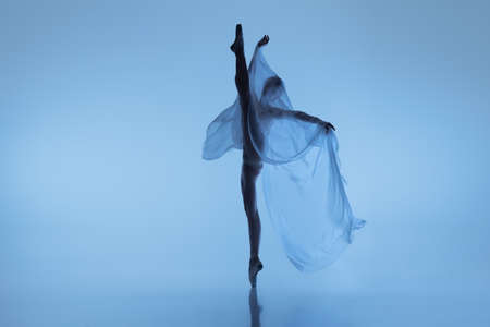 Portrait of flexible woman, graceful ballerina dancing with fabric, cloth isolated on blue studio background. Grace, art, beauty concept. Weightless, tenderness.の写真素材