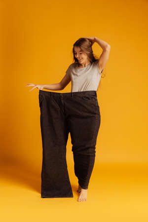Portrait of young slim woman standing in giant black pants isolated over yellow background. Weight loss conceptの写真素材