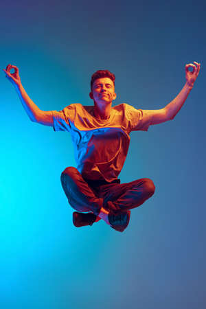 Portrait of young calm man, student sitting in lotus position in air isolated over blue studio...