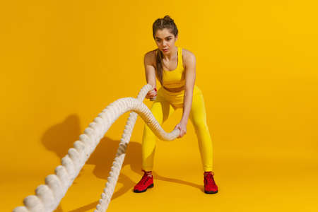 Studio shot of sportive slim girl workout with sports equipment isolated on bright yellow studio...