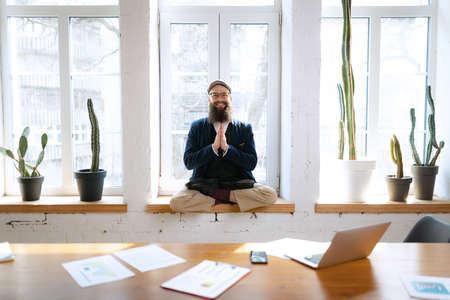 Young bearded man, office clerk having fun, doing yoga on wooden table in modern office at work time with gadgets. Concept of business, healthy lifestyle, sport, hobbyの写真素材