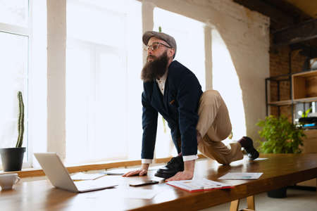 Young bearded man, office clerk having fun, doing yoga on wooden table in modern office at work time with gadgets. Concept of business, healthy lifestyle, sport, hobbyの写真素材