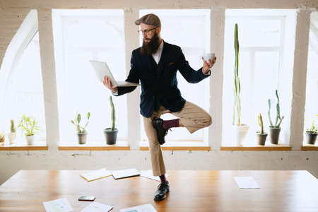 Young bearded man, office clerk having fun, doing yoga on wooden table in modern office at work time with gadgets. Concept of business, healthy lifestyle, sport, hobbyの写真素材