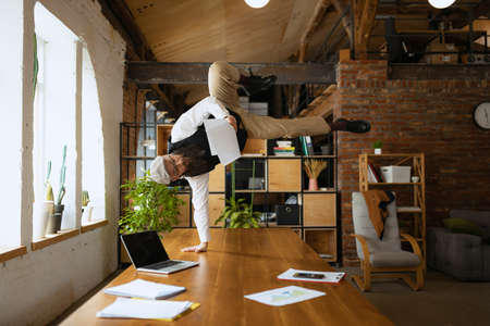 Young bearded man, office clerk having fun, doing yoga on wooden table in modern office at work time...