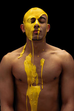 Young man being covered with thick yellow paint isolated over black background. Front viewの写真素材