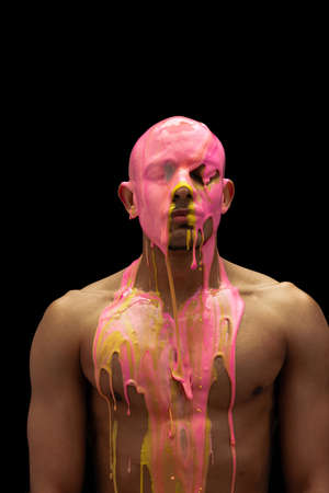 Portrait of young muscular man covered with thick pink and yellow paint isolated over black backgroundの写真素材